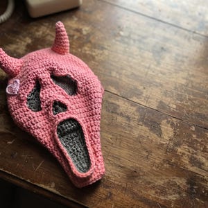 May include: A pink crocheted balaclava featuring two pointed horns on top and a ghost-like face design with dark gray eye and mouth openings. A small, light pink heart-shaped patch is stitched onto the side of the pink yarn head covering.