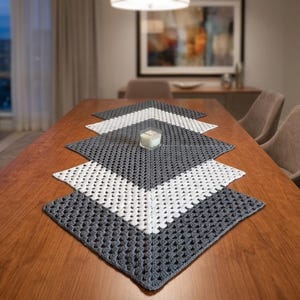 Könnte beinhalten: Handgefertigter Tischläufer mit geometrischem Muster. Der Läufer besteht aus gehäkeltem Garn in abwechselnden Abschnitten aus Weiß und Grau. Eine kleine Kerze in einem Glashalter steht auf dem Läufer, der auf einem Holztisch liegt.