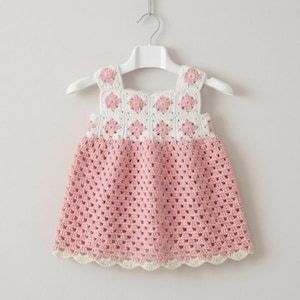 May include: A handmade, pink and white crochet dress for a baby or toddler, hanging on a white hanger. The dress features a square neckline and a scalloped hem. The top section has a white background with pink floral accents.