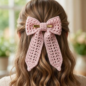 May include: A pale pink, crocheted hair bow with a gold-coloured clip. The bow is attached to the back of someone's head, with the long tails of the bow cascading down the hair. The bow has an open, lacy design.