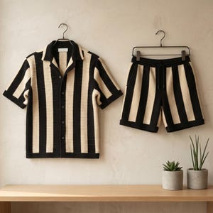 May include: A matching two-piece set of clothing, consisting of a short-sleeved button-up shirt and shorts, both with vertical black and cream stripes. The shirt has a collar and the shorts have a drawstring. The set is hanging on a wall.