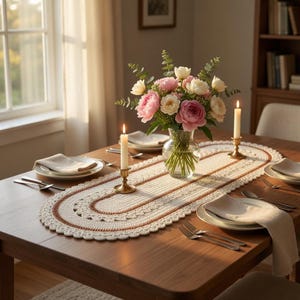 Puede incluir: Un camino de mesa ovalado de ganchillo con base color crema, anillos concéntricos de hilo marrón cobrizo y borde festoneado. El camino está sobre una mesa de madera con un jarrón de cristal con flores rosas y blancas y dos candelabros de latón.