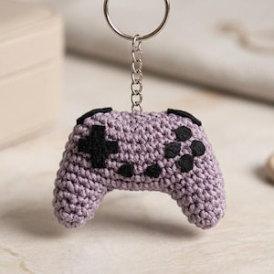 Peut inclure: Un porte-clés en forme de manette de jeu vidéo en crochet lavande avec des détails de boutons noirs. Le porte-clés est suspendu à une chaîne et un anneau argentés. L'arrière-plan comprend une boîte à bijoux beige et une lanière en cuir.