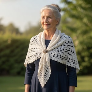May include: A woman wearing a cream-colored, crocheted shawl with a decorative brooch. The shawl is draped over a dark blue dress. The background is a sunny outdoor scene with green grass and trees, suggesting a spring or summer day.