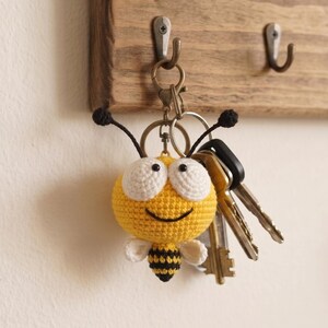 May include: A crocheted bee keychain featuring a round yellow head with large white eyes, a black stitched smile, and two black antennae. The body has yellow and black stripes with small white wings. It is attached to a metal ring with several keys.
