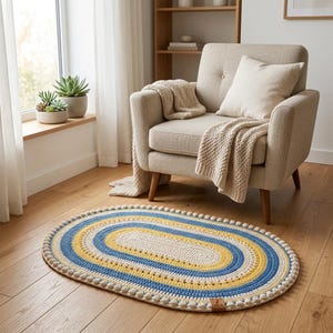 May include: An oval-shaped crochet rug featuring concentric bands of cream, yellow, and blue yarn. The rug is finished with a textured bobble-stitch border and includes a small rectangular brown leather tag with the text 7 MCA at the edge.