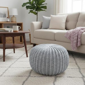 Op de afbeelding: Een woonkamer met een lichtgrijze gebreide poef, een beige bank met een lavendelkleurige deken en een donkerbruine ronde salontafel. De kamer heeft een neutraal kleurenpalet met een patroonkleed en houten planken.