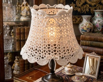 May include: A cream-colored crochet lampshade with a scalloped edge sits atop a dark bronze lamp base. The shade is intricately patterned, and a small bow adorns the front. The lamp is on a wooden table with a decorative rug.