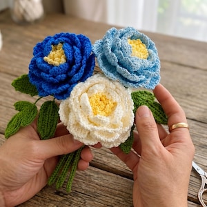 Puede incluir: Un ramo de flores de ganchillo hecho a mano con tres flores en tonos azules y crema, con centros amarillos. El ramo incluye hojas y tallos verdes, y lo sostiene una persona.