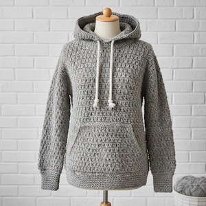 Könnte beinhalten: Ein grauer Häkel-Hoodie mit Kapuze und Vordertasche. Der Hoodie hat lange Ärmel und einen Kordelzug. Das Kleidungsstück wird auf einer Schaufensterpuppe vor einer weißen Backsteinmauer präsentiert.