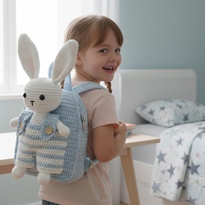 May include: A light blue crochet backpack with a white bunny attached. The bunny has long ears and is wearing blue and white striped overalls. The backpack is on a young girl's back.