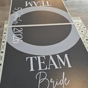 Könnte beinhalten: Ein schwarzer Beer-Pong-Tisch mit weißem Text, der Team Groom, Team Bride und 5 September 2026 liest. Die Tischoberfläche hat einen großen grauen Kreis in der Mitte und ist mit einer dreieckigen Anordnung von zehn schwarzen Plastikbechern.