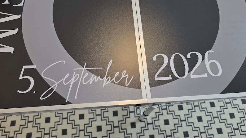 May include: A dark gray and black folding table surface featuring a circular design with the white text 5. September 2026 printed across the center. The table has a metallic silver frame and a visible seam running vertically through the middle.