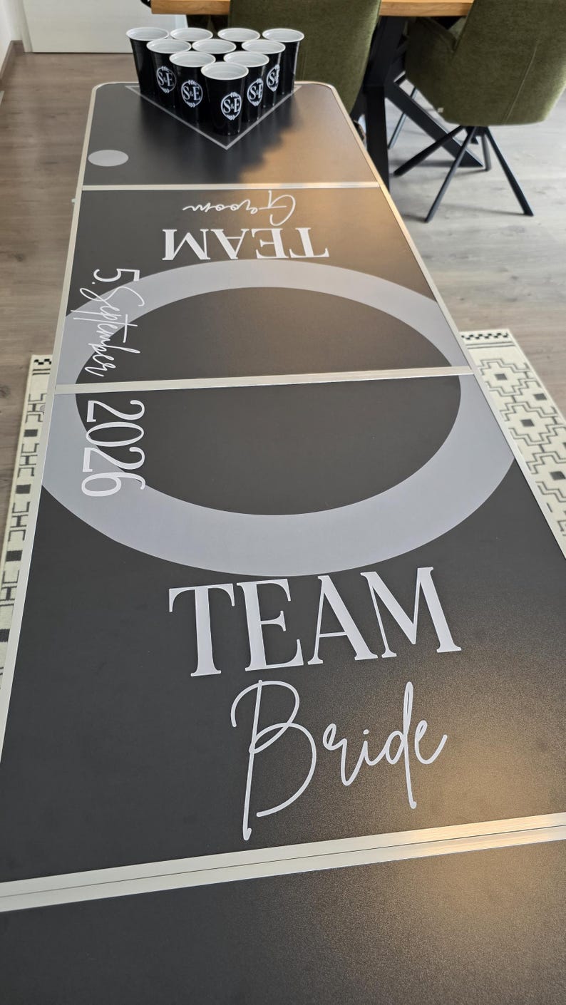 May include: A black beer pong table featuring white text that reads Team Groom, Team Bride, and 5 September 2026. The table surface has a large gray circle in the center and is topped with a triangular arrangement of ten black plastic cups with white logos.