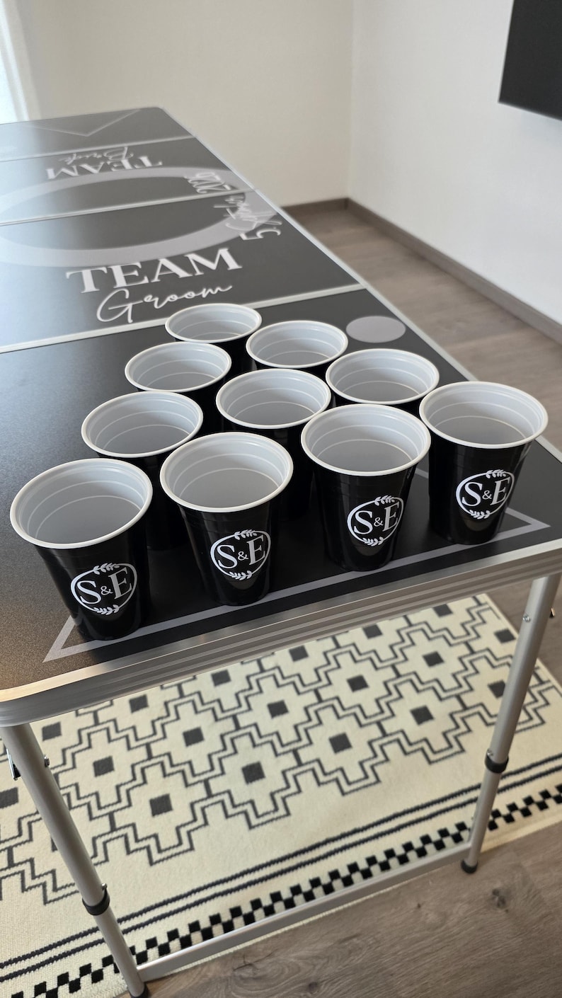 May include: A set of ten black plastic cups arranged in a triangle on a black game table. Each cup features a white circular logo with the letters S&E surrounded by a leaf design. The table surface displays the text TEAM Groom and 5 Sept 2026.