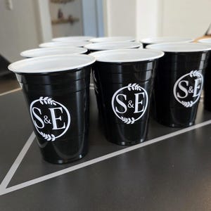 May include: A collection of black plastic party cups with a white interior rim. Each cup features a circular white logo on the front containing the letters S&E surrounded by a decorative leaf wreath design. The cups are arranged in a triangular formation.