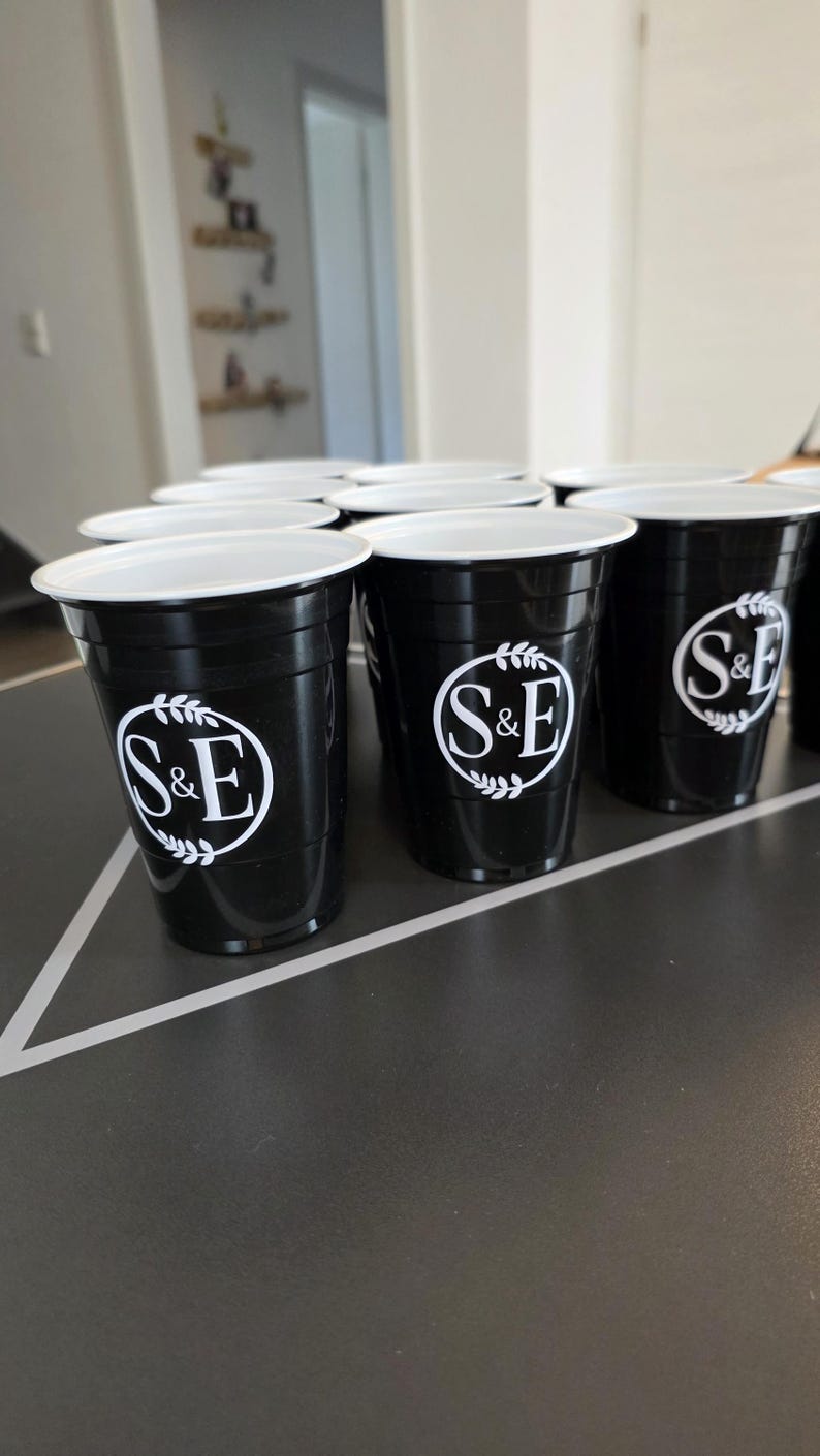 May include: A collection of black plastic party cups with a white interior rim. Each cup features a circular white logo on the front containing the letters S&E surrounded by a decorative leaf wreath design. The cups are arranged in a triangular formation.