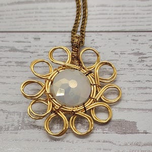 May include: A gold-colored wire-wrapped pendant necklace. The pendant features a large, faceted, off-white gemstone at the center, surrounded by a floral-like design. The chain is also gold-colored.