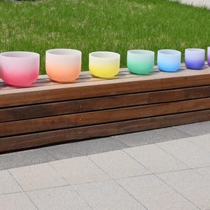 May include: A collection of seven frosted crystal singing bowls in a gradient of colors, from pink to purple, arranged on a wooden bench. Two purple carrying cases are on either end. The bowls vary in size.