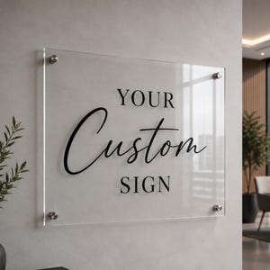 May include: A clear acrylic sign with the text "YOUR Custom SIGN" in black. The sign is mounted on a light grey wall with silver-coloured hardware. The sign is rectangular and appears to be in an office setting.