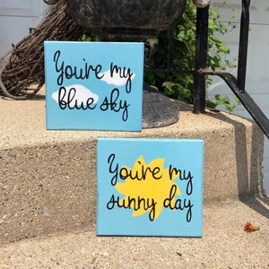 May include: Two blue painted wooden signs with white clouds and a yellow sun. The top sign says "You're my blue sky" and the bottom sign says "You're my sunny day".