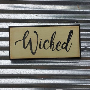 May include: A wooden sign with a black frame and a cream background. The word "Wicked" is written in black cursive lettering.