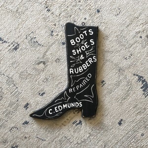 May include: Black wooden boot-shaped sign with white lettering that reads "Boots Shoes & Rubbers Repaired C. Edmunds".
