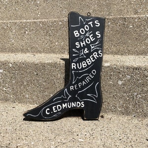 May include: Black wooden boot-shaped sign with white lettering that reads "Boots Shoes & Rubbers Repaired C. Edmunds".