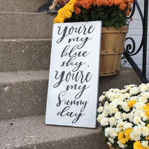 May include: White wooden sign with black calligraphy that reads "You're my blue sky, You're my sunny day". The sign is on a set of concrete steps with a potted plant of yellow and orange flowers to the right.