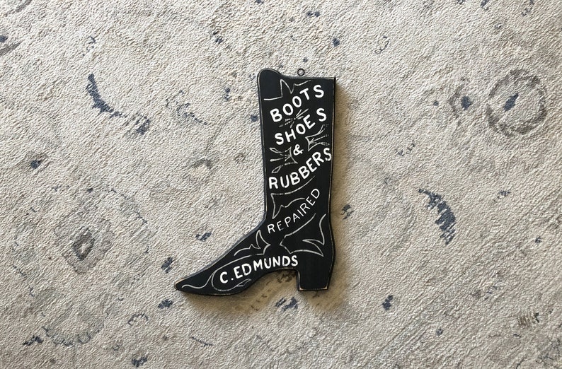 May include: Black wooden boot-shaped sign with white lettering that reads "Boots Shoes & Rubbers Repaired C. Edmunds".