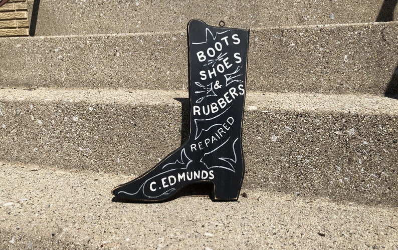 May include: Black wooden boot-shaped sign with white lettering that reads "Boots Shoes & Rubbers Repaired C. Edmunds".