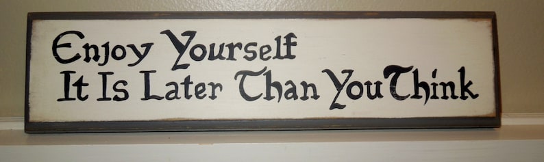 K&ouml;nnte beinhalten: Ein wei&szlig;es Holzschild mit schwarzer Beschriftung, auf dem "Enjoy Yourself It Is Later Than You Think" steht.