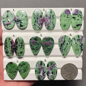 Op de afbeelding: Een verzameling van zestien gepolijste zoisiet en robijn cabochons, gerangschikt in paren op een wit displayplateau en genummerd van één tot acht. De stenen hebben een gevlekte groene basis met zwarte insluitingen en magenta-roze robijnvlekken.