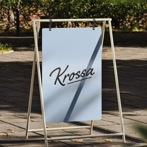 May include: A light blue rectangular sign suspended from a cream-colored metal A-frame stand. The sign features the word Krossa written in black cursive script with a horizontal line underneath. The structure is held by two dark leather straps.