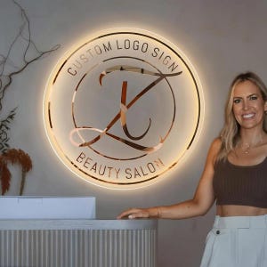 May include: A circular, wall-mounted, illuminated sign featuring a metallic rose gold finish. The sign displays the text CUSTOM LOGO SIGN at the top and BEAUTY SALON at the bottom, with a large, stylised letter J in the centre, all backlit by a warm glow.