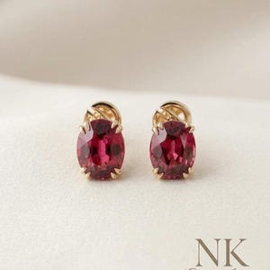 Puede incluir: Un par de pendientes de oro con tachuelas, cada uno con una gran piedra preciosa roja oscura de corte ovalado. Los pendientes están engastados en metal dorado con un diseño texturizado alrededor de la piedra preciosa. El fondo es una tela suave blanquecina.