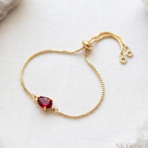 May include: A gold-colored adjustable bracelet featuring a teardrop-shaped, deep red gemstone. The delicate chain has a box link design and a sliding bead closure. The bracelet is displayed on a white surface.