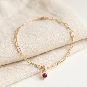 Puede incluir: Una pulsera de cadena dorada con un pequeño colgante de piedra preciosa roja en forma de corazón. La pulsera está hecha de eslabones ovalados y se muestra sobre una tela beige. El colgante es de color rojo intenso.