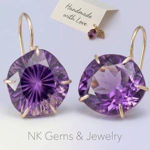 May include: A pair of gold-colored earrings with large, faceted, purple amethyst stones. The earrings have a hook design and are set with gold prongs. A small tag reads "Handmade with Love."