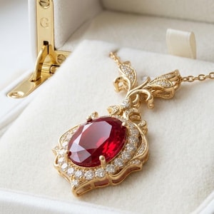 May include: A gold pendant necklace with a large oval ruby gemstone at the center, surrounded by small diamonds. The pendant has an ornate, filigree design. The necklace is displayed in a white jewelry box with gold hardware.