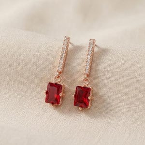 May include: A pair of rose gold earrings with rectangular, deep red gemstones. The top portion of each earring features a slender bar adorned with small, clear crystals. The earrings are displayed on a neutral-toned fabric.