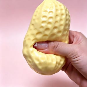 May include: A yellow peanut-shaped stress ball with a textured surface is being squeezed by a hand. The background is a solid pink color. The stress ball is a light yellow color.