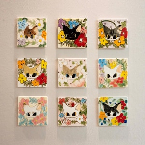 Puede incluir: Un conjunto de nueve azulejos de cerámica cuadrados dispuestos en cuadrícula, cada uno con una ilustración pintada a mano de una cara de gato rodeada de flores coloridas. Los gatos varían en color, incluyendo patrones negros, blancos y atigrados.
