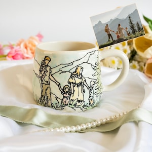 May include: A ceramic mug featuring a hand-drawn black ink illustration of a family of four walking in a mountainous landscape, with soft watercolour accents in pale yellow and blue, matching the photograph displayed above the mug.