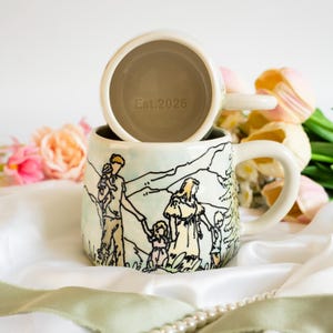 Peut inclure: Deux tasses en céramique avec une base de couleur crème. La tasse inférieure présente une illustration dessinée à la main d'une famille de quatre personnes dans un paysage. La tasse supérieure montre l'intérieur avec le texte Est.2026 imprimé.