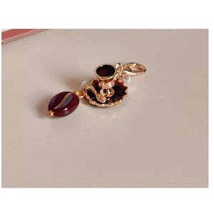 以下が含まれることがあります： ミニチュアのコーヒーカップとソーサー、濃い赤色のコーヒー豆のアクセントが付いたゴールドトーンのチャーム。チャームには、簡単に取り付けられるロブスタークラスプが付いています。コーヒーカップとソーサーは黒のエナメルで詳細に描かれています。