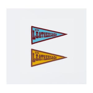 May include: Two pennant-shaped patches. The top patch is light blue with maroon trim and the text "T.S.L. LEATHERS & CO" in white. The bottom patch is yellow with brown trim and the same text.