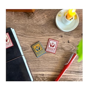 May include: Two small, rectangular patches with the word "FRAGILE" and "HANDLE WITH LOVE" printed on them. One patch is red with white text and the other is yellow with blue text. A red pen, a notebook, and a candle are also visible.