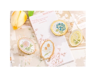 May include: Four gold-toned enamel pins featuring floral designs. The pins are in the shape of oval frames with white backgrounds and gold borders. The flowers include a pink rose, a cluster of white flowers, a blue hydrangea, and a cluster of yellow flowers.