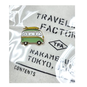 Op de afbeelding: Een groene en witte emaille pin met een vintage busje met een surfboard erop. De pin heeft een gele omtrek en de tekst "TRAVEL FACTOR" en "TFA" zijn zichtbaar op de achtergrond.